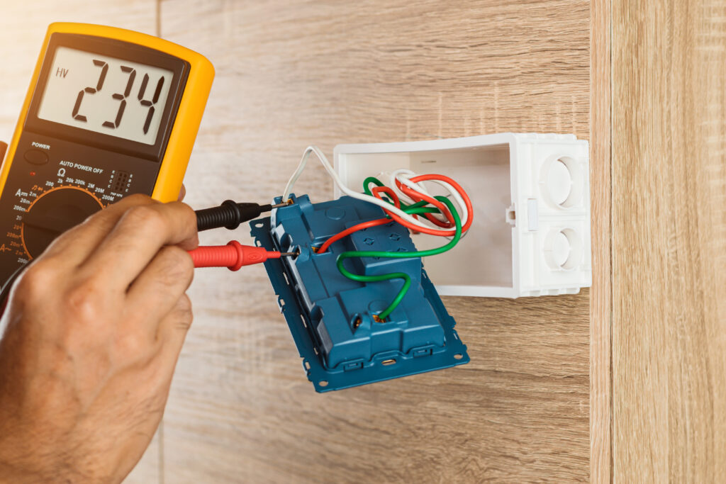 Electrician using a digital meter to measure the voltage at a wall socket on a wooden wall.